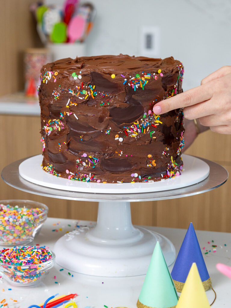 image of sprinkles being pressed gently into the side of a yellow cake that's been frosted with chocolate buttercream