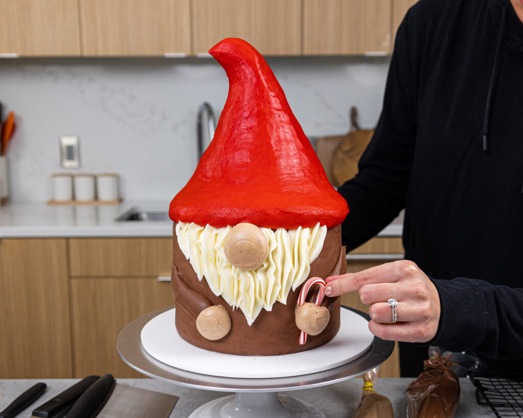 image of chelsey white of chelsweets adding a mini candy cane into the hand of an adorable gnome cake she made with vanilla cake layers, buttercream and rice krispies