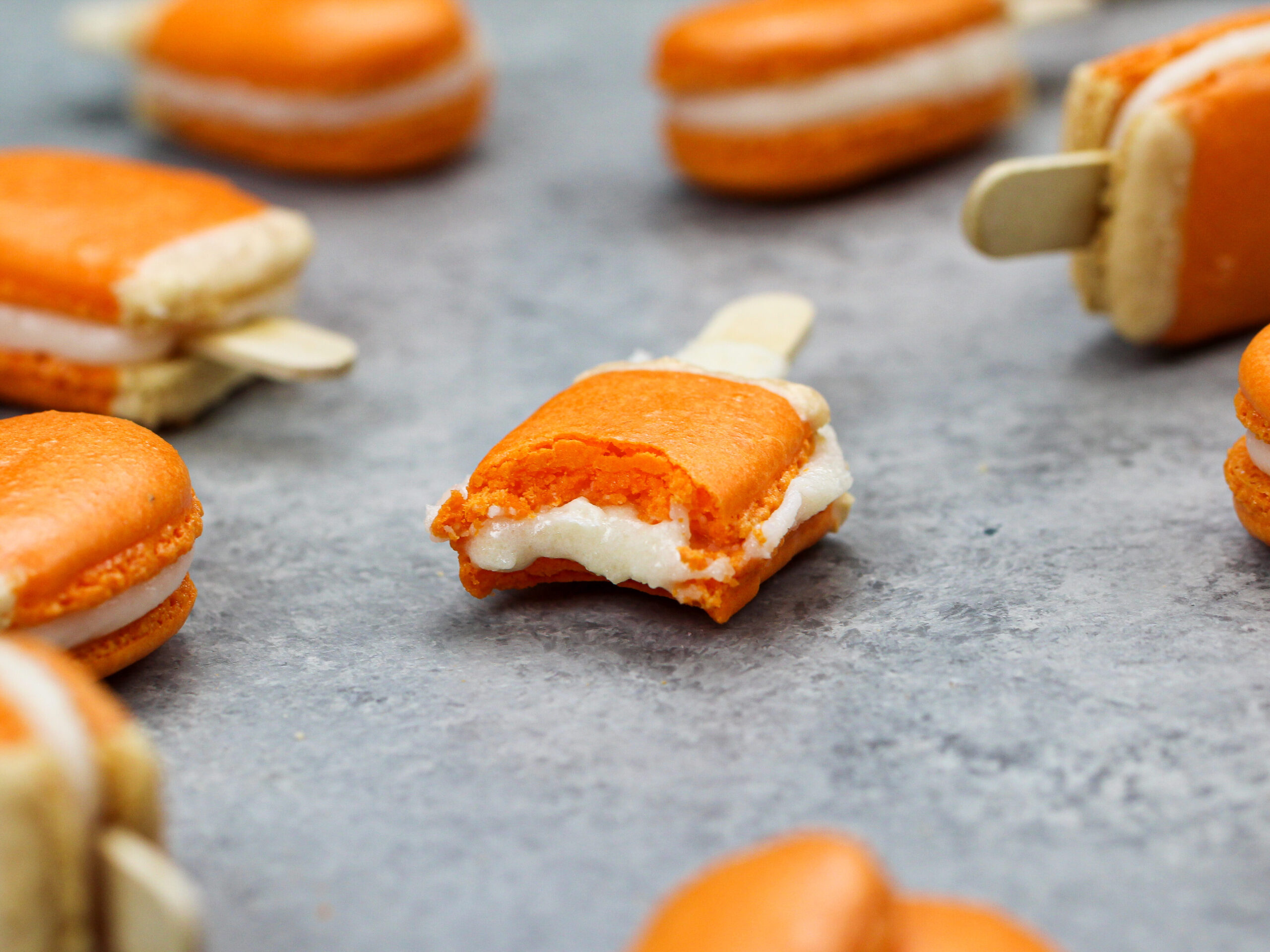 image of mini orange creamsicle macarons filled with orange creamsicle buttercream that have been bitten into to show the filling and full shells