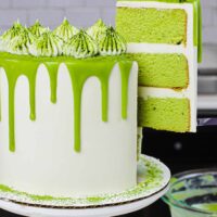 image of matcha cake slice on a plate with pretty matcha drip cake in background