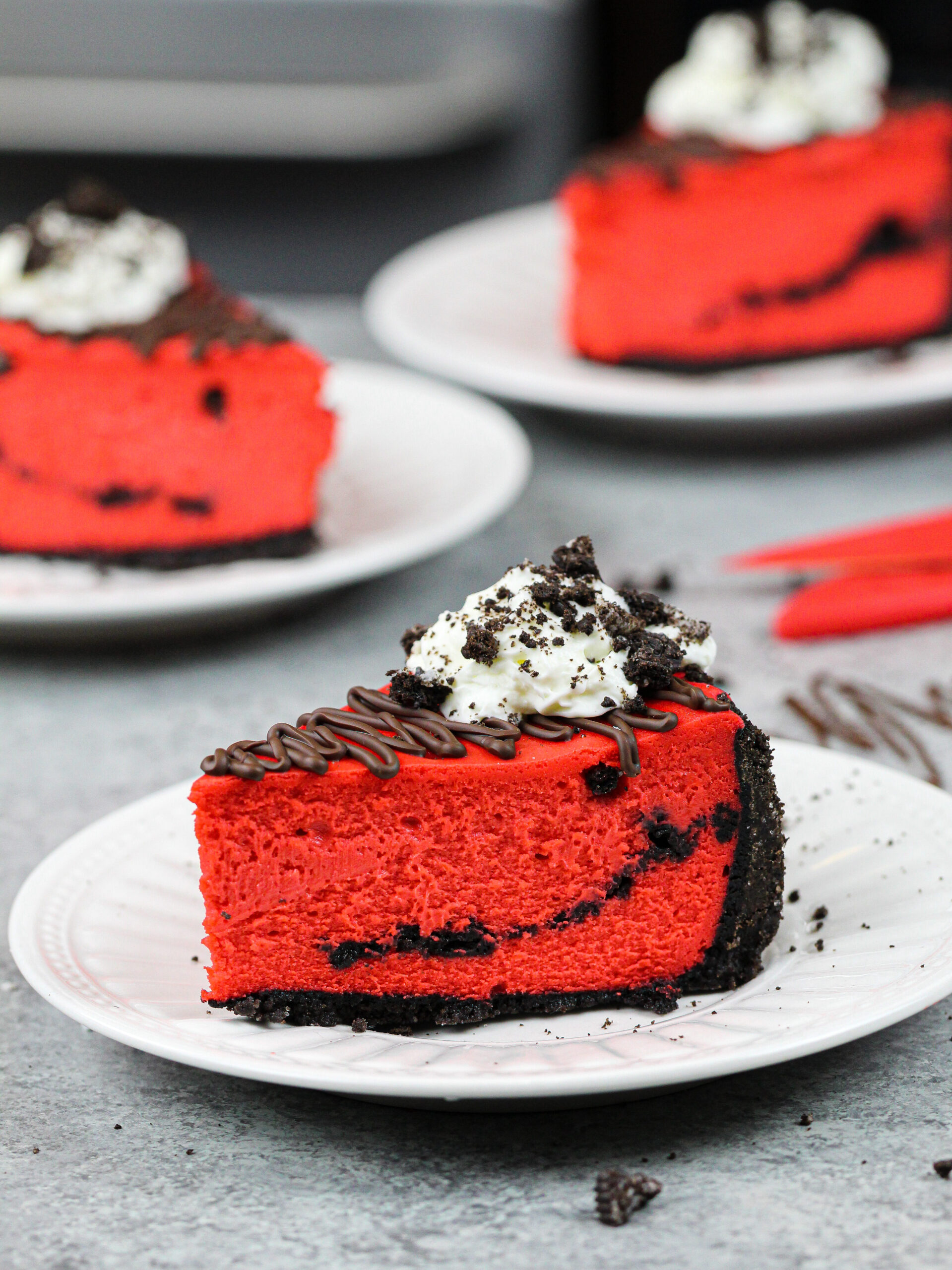 image of a slice of red velvet oreo cheesecake decorated with melted chocolate, whipped cream and crushed oreos