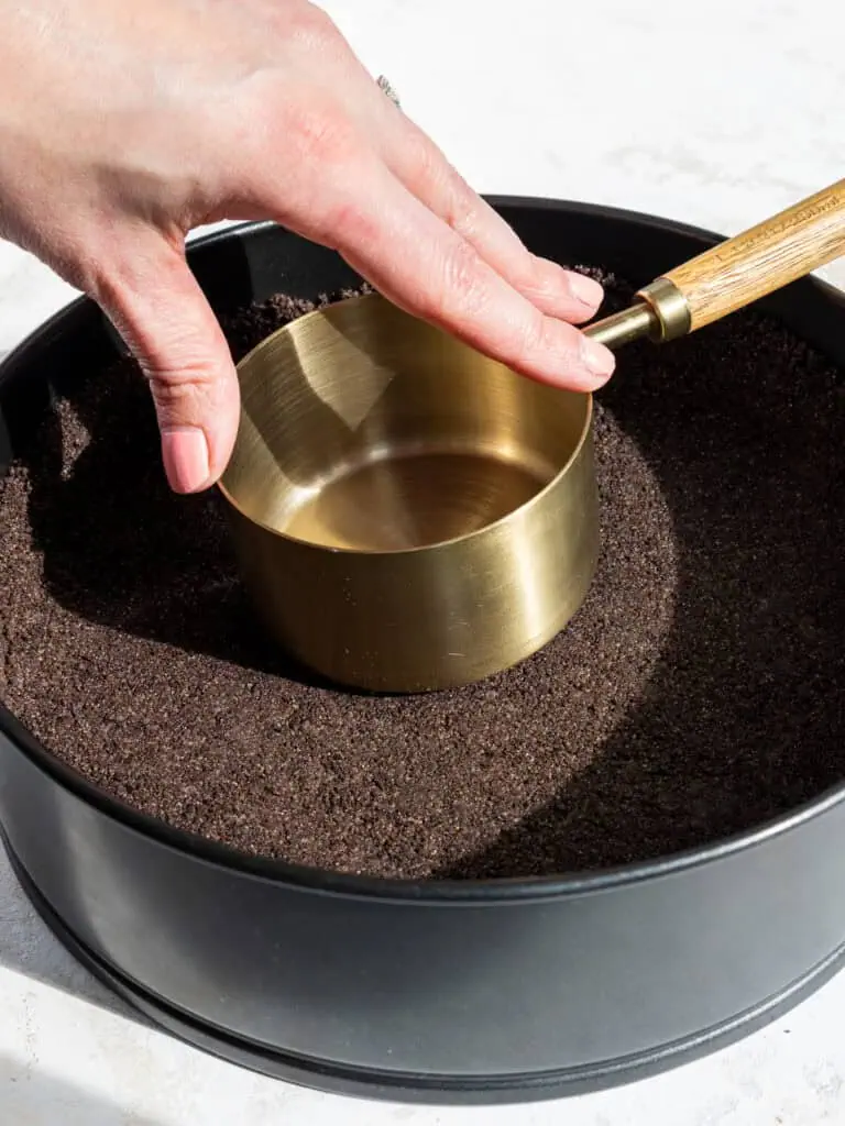 image of an oreo cookie crust being pressed into a cheesecake pan with a measuring cup