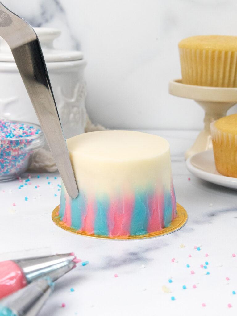 image of pink and blue frosting being spread on a mini cake with a small offset spatula