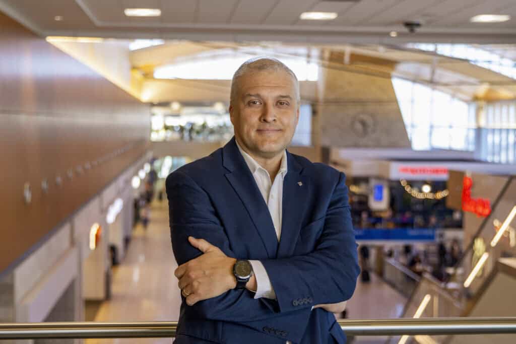 Photo of executive is a blue jacket with his arms crossed and interior of airport terminal in the background.