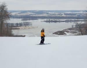 Lone woman at the top of a hill on skis overlooking a frozen river.