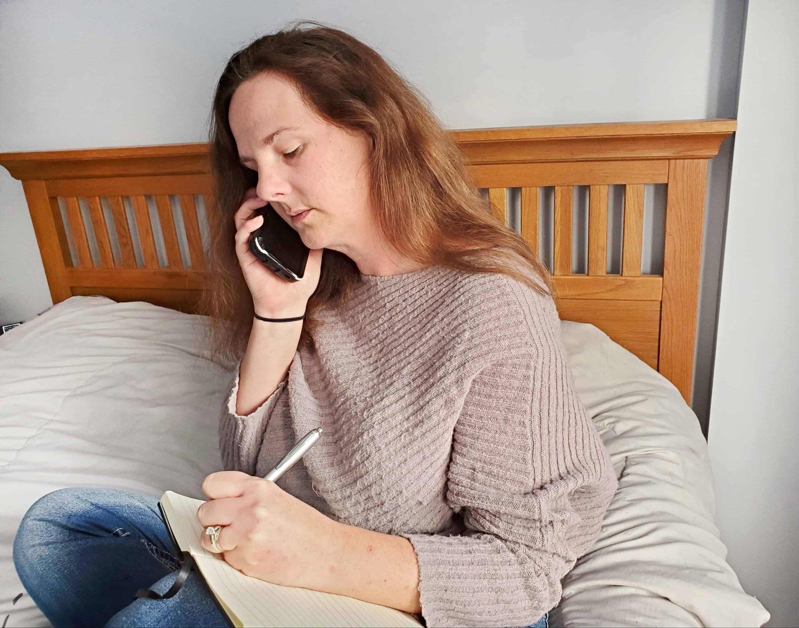 Woman taking notes while on a phone and sitting on the bed.