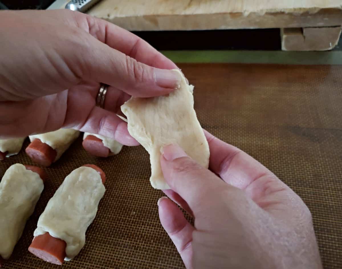 Two hands stretching dough into a rectangle over a partially filled tray of bagel dogs.