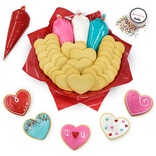 Heart shaped sugar cookies in a red tray with bags of colored icing around it and five decorated cookies below.