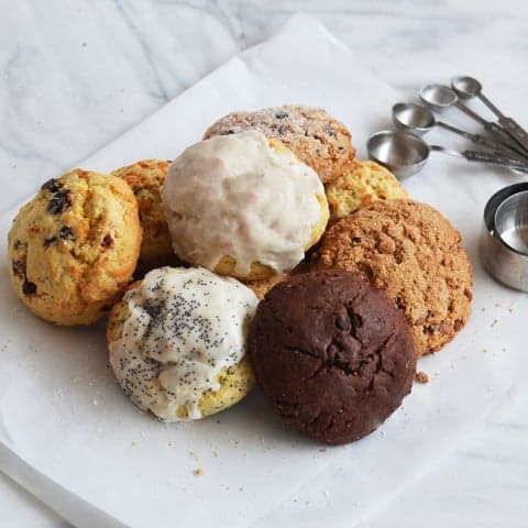 Variety of seven scones sitting on a white background with measuring spoons in the background.