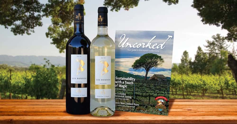 Two wine bottles on a wooden slab with a winery in the background and Uncorked pamphlet next to them.