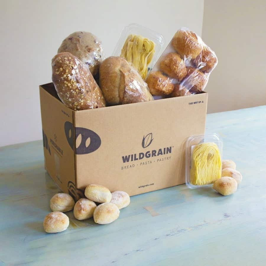 Wildgrain subscription box on a blue table with bread loaves, pasta, and rolls in and round the box.