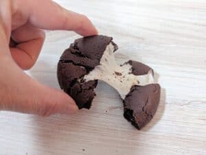 Hand pulling up half a gooey chocolate marshmallow cookie while it rests on a wooden board.