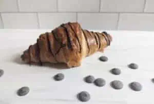 Chocolate drizzled croissant on a white background with chips around it.