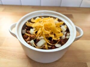 Image shows slow cooker pork chili served in a serving pot with toppings.