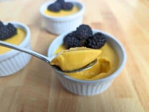 Image shows spoonful of easy to make mango mousse with two mango mousse in a ramekin in the background.