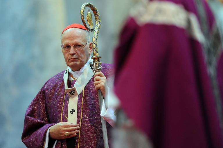 Um cardeal está em uma cerimônia religiosa, vestindo uma túnica roxa e segurando um cajado. Ele tem uma expressão séria e usa óculos. Ao fundo, outras figuras religiosas também estão presentes, vestindo vestes semelhantes.