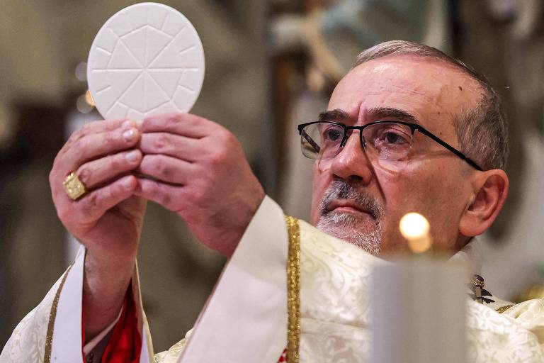 Um sacerdote, com cabelo grisalho e óculos, está segurando uma grande hóstia branca em suas mãos, enquanto a levanta em direção ao alto. Ele está vestido com uma vestimenta litúrgica dourada e vermelha. Ao fundo, há uma iluminação suave e elementos decorativos de uma igreja.