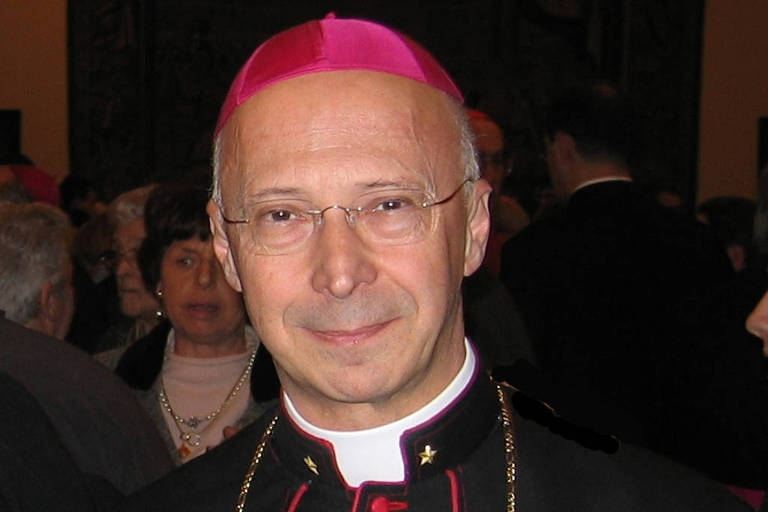 Um homem vestido com vestes eclesiásticas, incluindo uma batina preta com detalhes em rosa e um chapéu rosa. Ele usa óculos e tem cabelo claro. Ao fundo, há pessoas em um ambiente social.