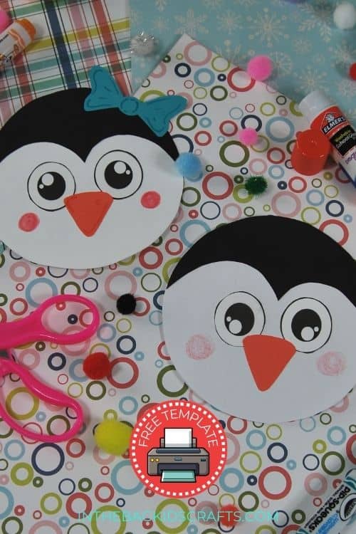 A colorful penguin craft setup featuring two round penguin cards made from a free printable, surrounded by craft supplies like scissors, glue, markers, and pom-poms on patterned paper.