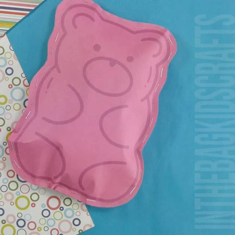 A pink candy gummy bear craft made from paper, stitched around the edges, displayed on a blue background with patterned scrapbook paper, making it a fun paper craft for kids.