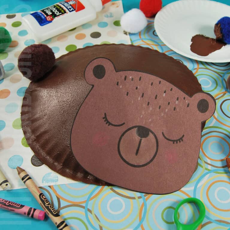An adorable bear craft for kids made from a painted paper plate, featuring a hibernating animal design, surrounded by craft supplies like crayons, glue, and pom-poms.