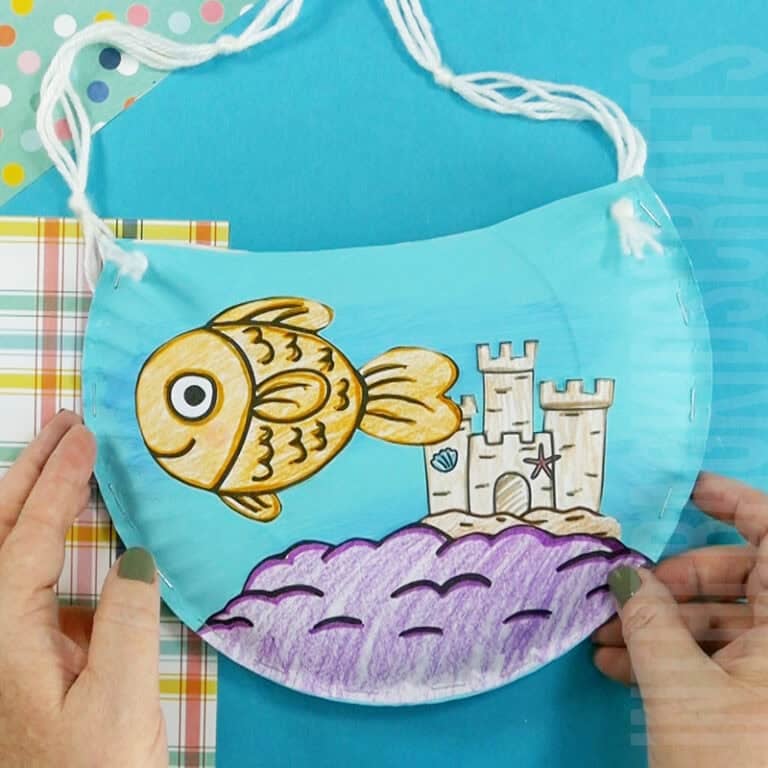 A colorful fish bowl craft for kids made from a paper plate craft, featuring a hand-colored goldfish craft swimming near a sandcastle with string handles for hanging.