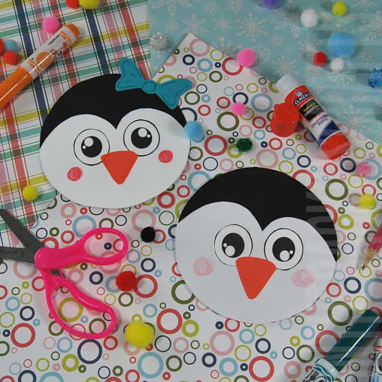 A colorful penguin craft setup featuring two round penguin cards made from a free printable, surrounded by craft supplies like scissors, glue, markers, and pom-poms on patterned paper.