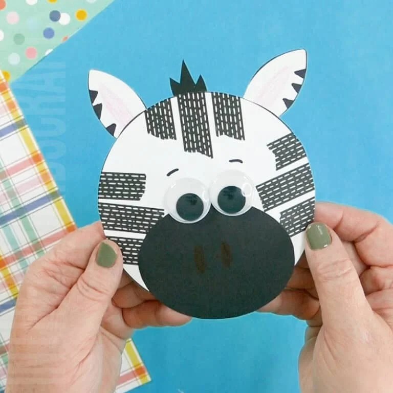 A person holding a completed preschool zebra craft, a fun fine motor activity featuring a round paper zebra face with black stripes, googly eyes, and a blue background.