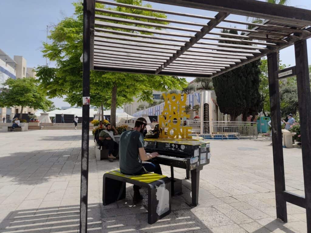 צעיר יושב לבדו ומנגן בפסנתר ציבורי בכיכר בירושלים, תחת שלט “You are not alone”