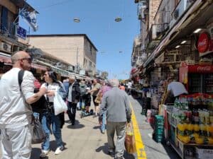 שוק מחנה יהודה הומה אדם ביום חמישי בצהריים, עם שמש חזקה ודגלי ישראל
