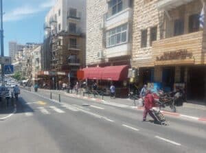 רחוב אגריפס בירושלים, צהריים חמים, אישה חוצה מעבר חצייה מול חזיתות מסחריות ובתי קפה