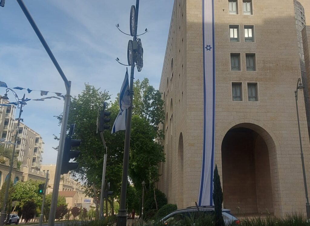דגל ישראל ענק תלוי על בניין באזור ממילא בירושלים, ברקע רחוב ודגלים נוספים
