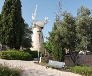 טחנת הרוח מונטיפיורי ההיסטורית במשכנות שאננים בירושלים, עם עצים וספסל ביום בהיר. סמל לבנייה היהודית מחוץ לחומות.