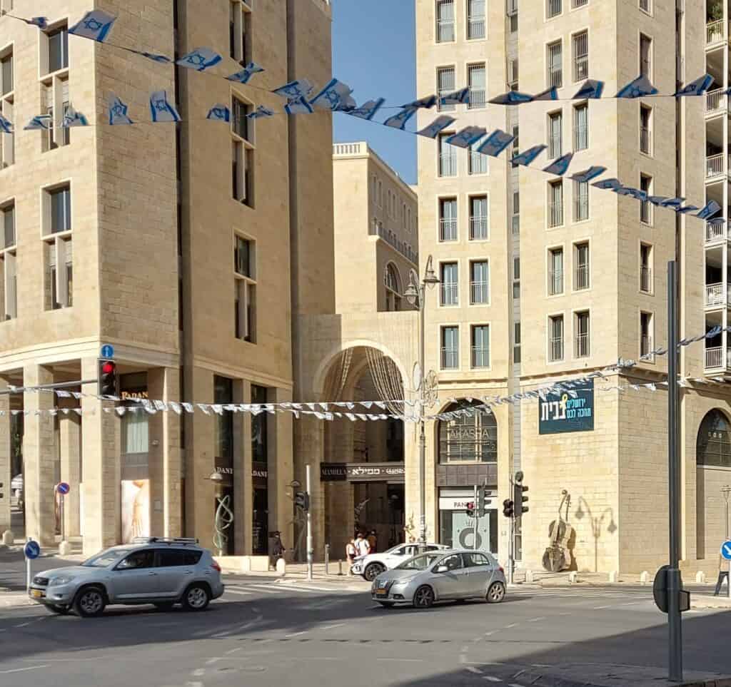 רחוב שוקק חיים סמוך לשדרות ממילא בירושלים ביום שמשי בהיר, עם אנשים הולכים, חנויות ומבנים מודרניים. משקף אווירת סופש בעיר.