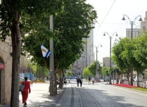 רחוב יפו בירושלים ביום שמשי בהיר, עם אנשים הולכים על המדרכה, עצים, בניינים ומסילת רכבת קלה.
