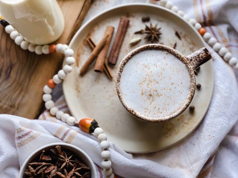 a chai tea latte sits on a plate filled with cinnimon sticks, star anise pods, whole cloves and all spice berries. With a bottle of milk near by