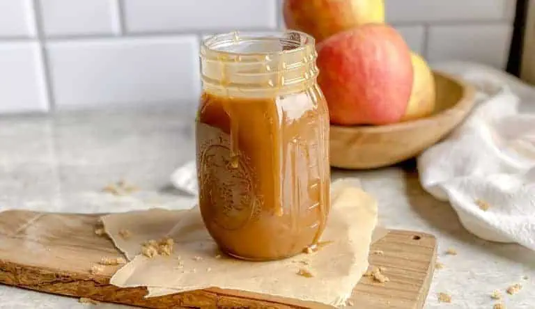 a jar of homemade caramel sauce on wooden cutting board with a bowl of red apples behind it.