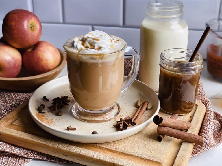a mug of apple cider chai tea latte sits on a tan plate with a jar of milk and caramel near by and red apples in background