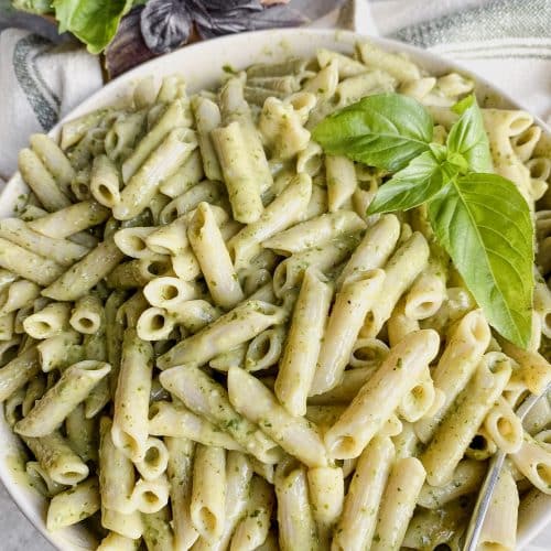 large bowl of gluten free penne pasta with dairy free pesto pasta sauce and fresh basil leaves on top