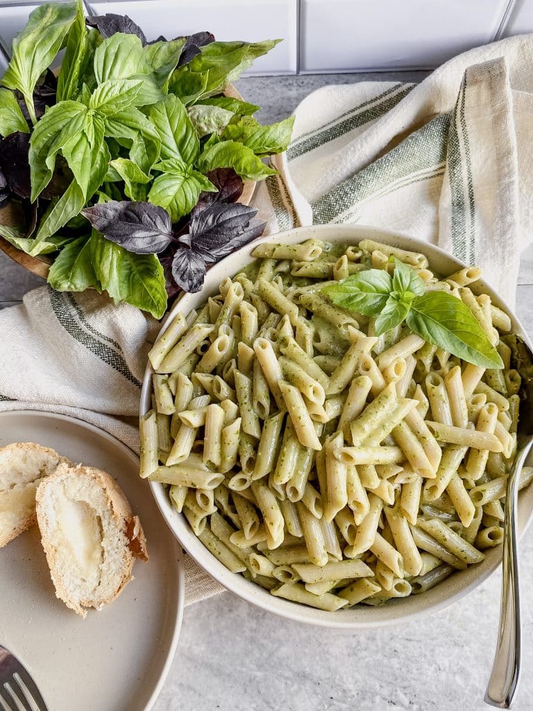 large bowl full of gluten free pesto pasta with creamy sauce and a bowl of fresh basil