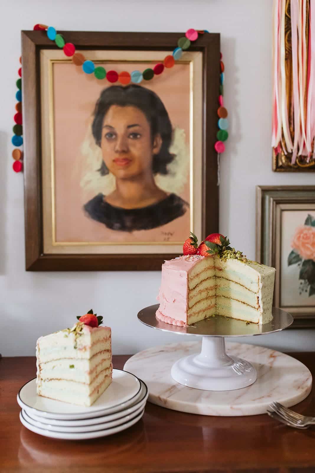 Slice of doberge cake and whole cake on a buffet with pictures behind.