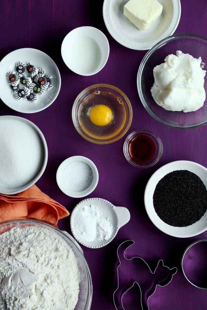 Ingredients for Halloween sugar cookie recipe in small bowls on purple board.