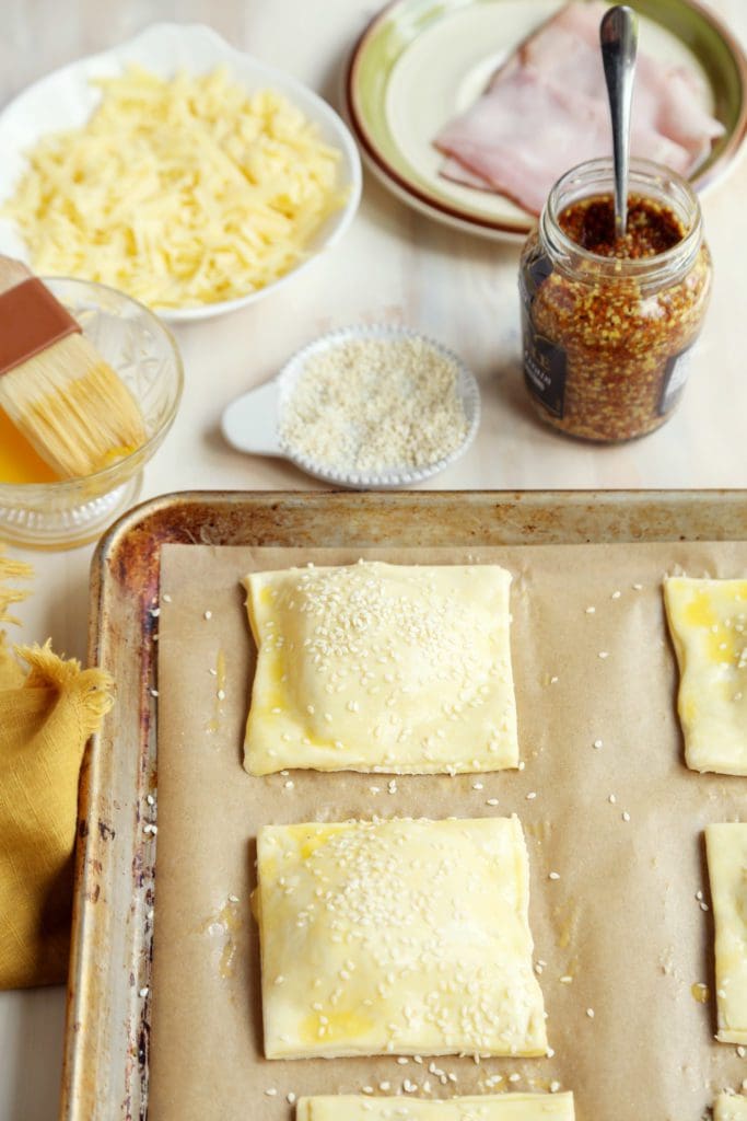 Ham and cheese croissant squares on parchment lined baking sheet with egg washed and sprinkled sesame seeds.
