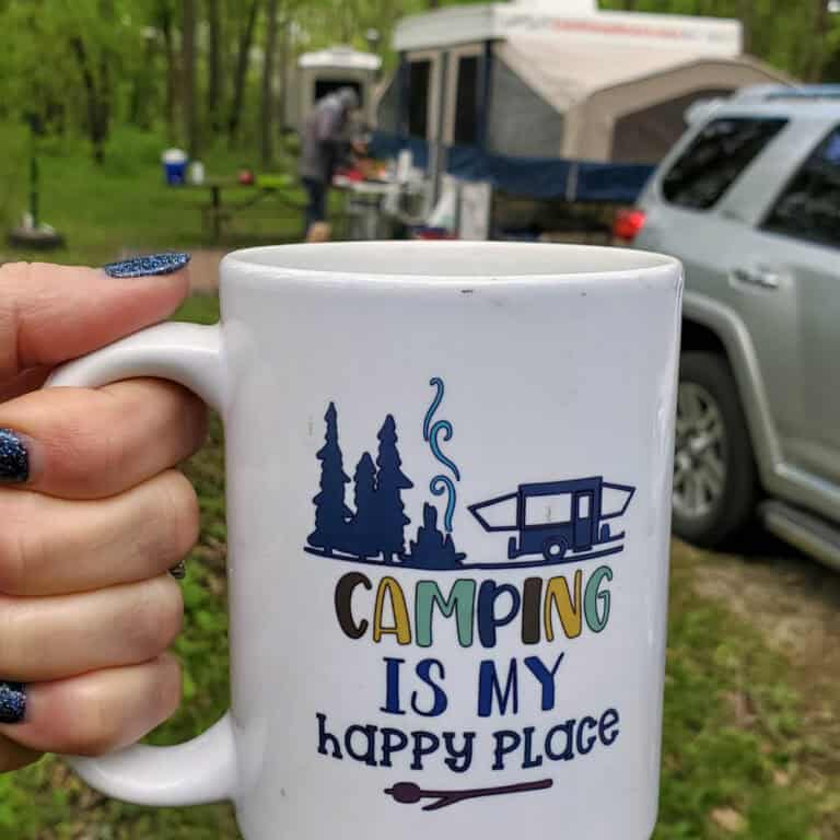 person holding a mug that says camping is my happy place at a campground