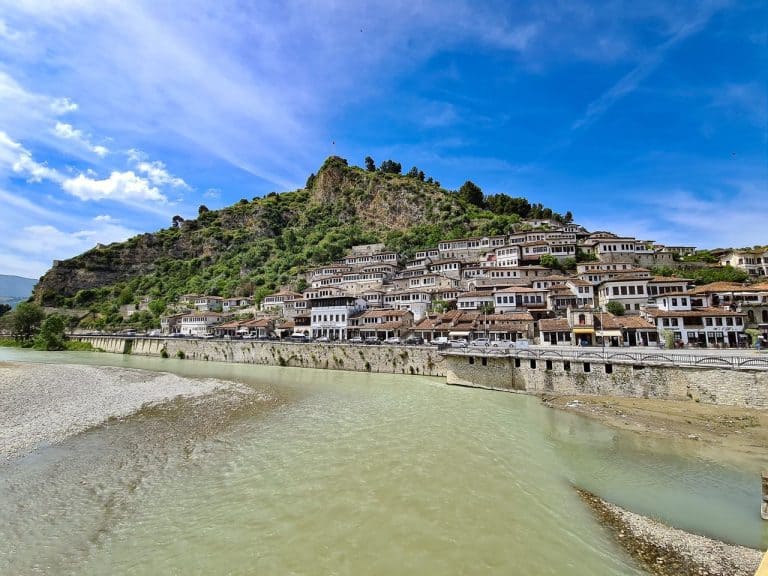 Panoramic view of Berat, Albania, featuring the iconic Ottoman-style houses in the Mangalem Quarter with their 'floating windows,' set against a lush hillside and the Osum River under a bright blue sky.