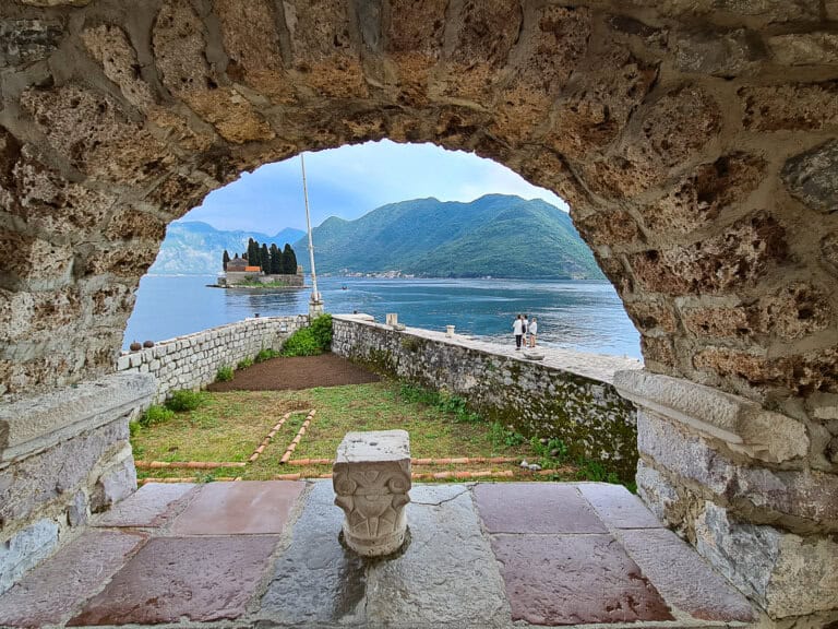 View of the island of St. George from Our Lady of the Rocks in Perast, Montenegro, framed by an ancient stone archway. A must-visit Instagrammable spot in the Bay of Kotor.
