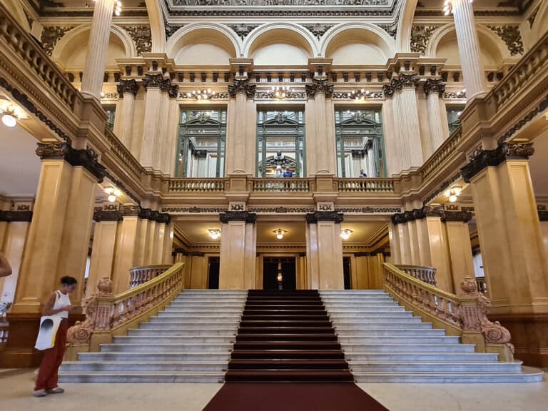 The grand staircase inside the Teatro Colon Buenos Aires. The opulent interior features marble steps, flanked by ornate balustrades and classical Corinthian columns. The ceiling boasts elegant stained glass and detailed plasterwork.