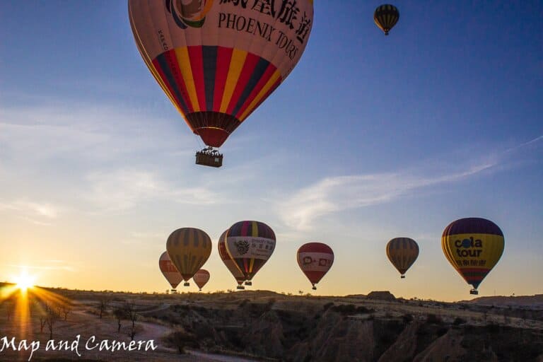 A breathtaking sunrise scene with multiple colorful hot air balloons floating in the sky. The background features a serene landscape with gentle hills, and the rising sun casts a warm glow on the horizon, emphasizing the silhouettes of the balloons. The photo is taken from one of the best cave hotels with view in Cappadocia