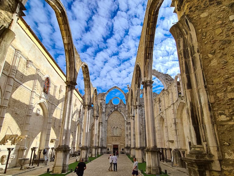 A wide-angle view of the Carmo Convent ruins in Lisbon, showing the open sky through the missing roof. The Gothic arches are still standing, framing the blue sky with fluffy white clouds. Visitors walk along the open-air museum, observing the historical remnants. The combination of ancient stone walls and arches with the vibrant sky creates a blend of history and natural beauty.
