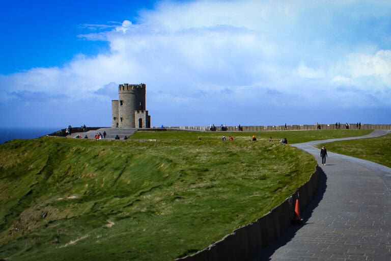 The image captures a scene at the Cliffs of Moher, featuring a robust, square stone tower standing prominently on the edge of the cliffs. The tower, with its battlements, serves as a viewing point for the many visitors visible in the distance. A paved walkway leads to the tower, and a visitor is seen walking towards it. The grass-covered landscape is typical of the rugged Irish coastline, with the grass appearing windswept, hinting at the strong breezes common to the area. The sky is bright with a mix of blue and scattered white clouds, suggesting a clear, fresh day. This setting is iconic for its natural beauty and the sense of history it conveys.